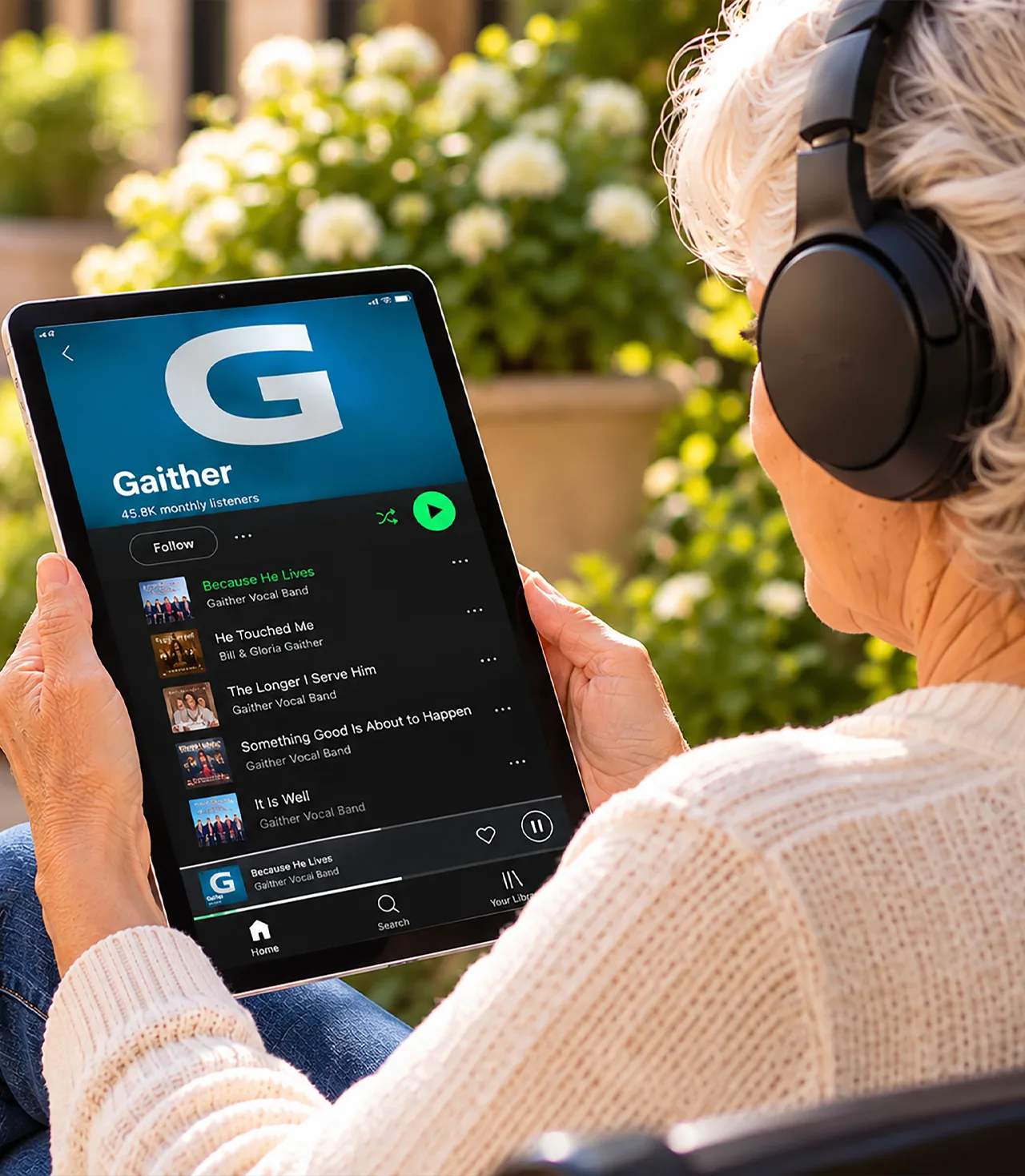 A woman with gray hair sitting on a bench outside, holding a tablet displaying a music app.